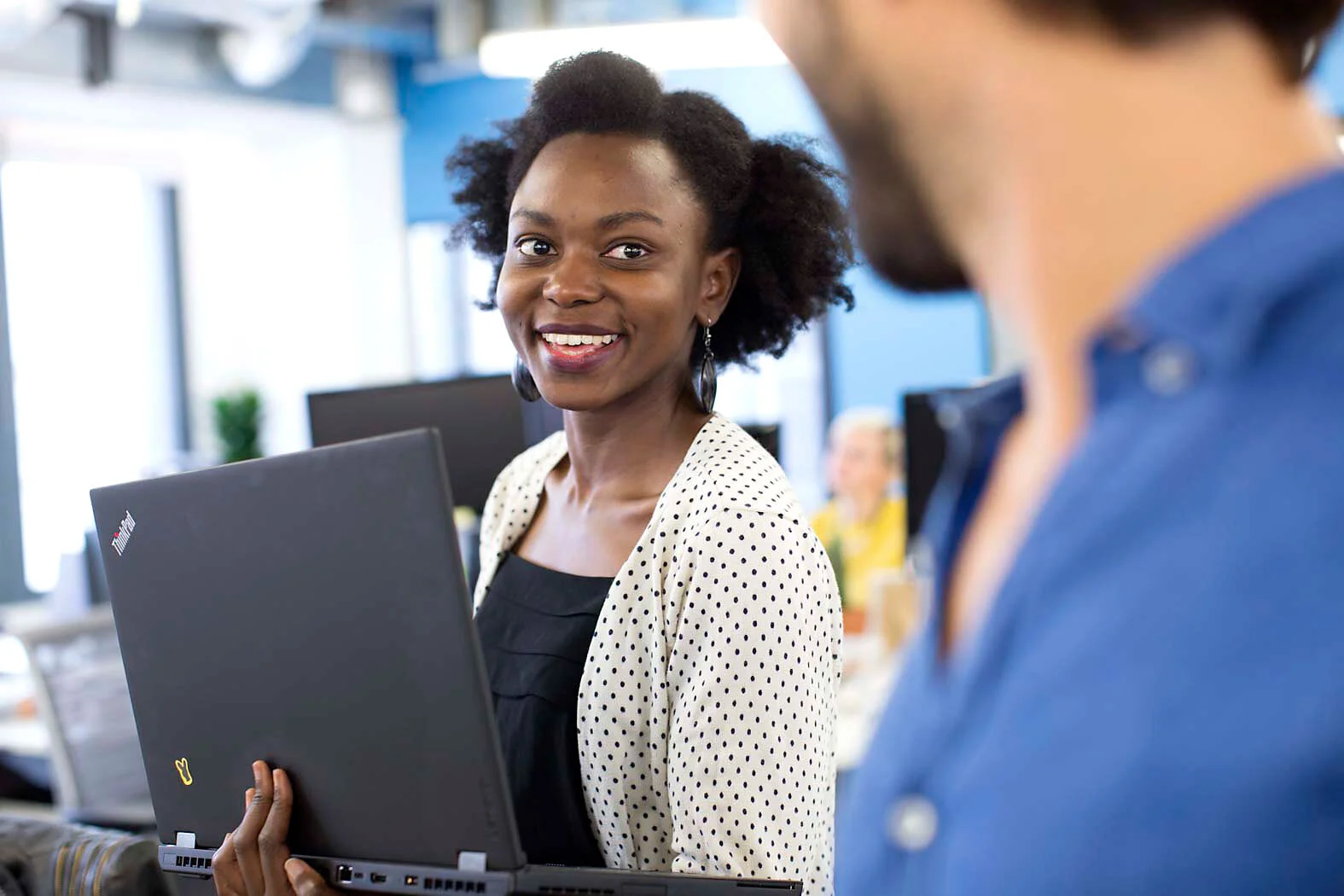 Woman holding a laptop in an office, smiling at a colleague.