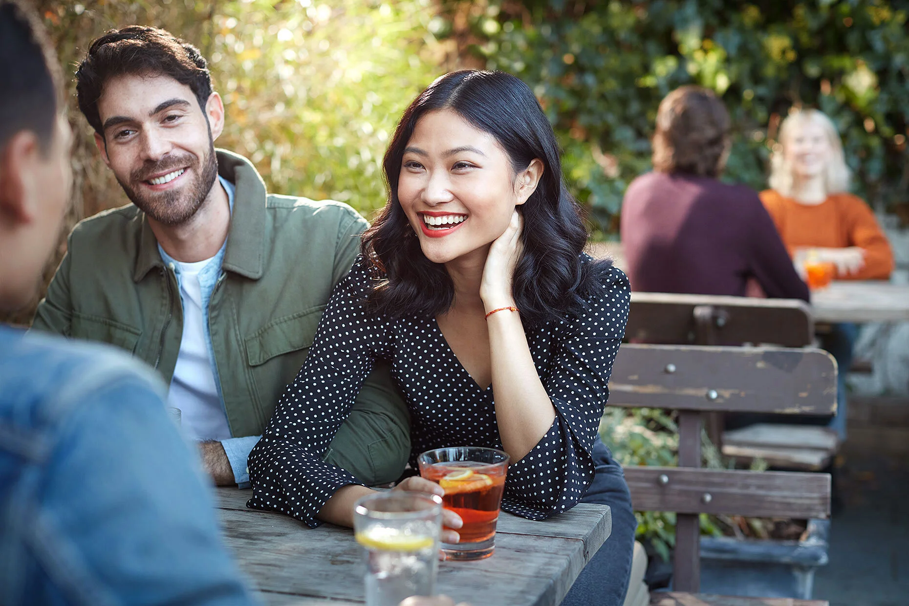 Group of people sitting outdoors at a wooden table, smiling and enjoying drinks.