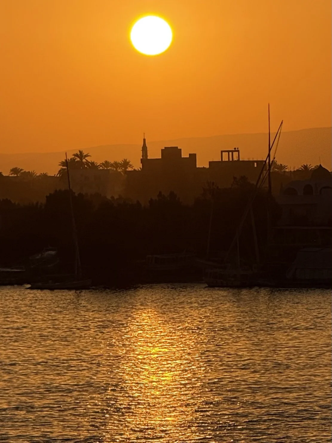 Sunrise over the Nile river with boats docked, silhouettes of buildings and trees, with a warm orange sky.