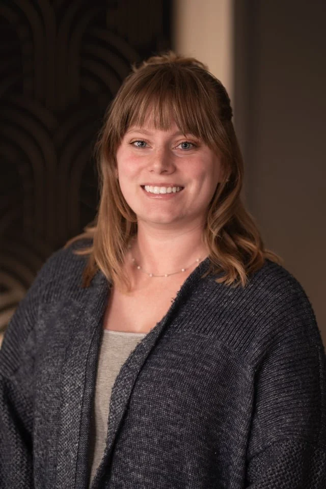 Tory Nicholas wearing a dark gray blazer over a light-colored top, standing indoors with a textured wall in the background.