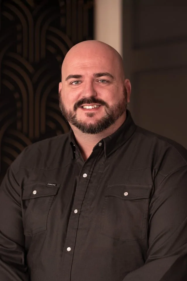 Rob Johnson wearing a black collared shirt, standing indoors with a patterned wall in the background.