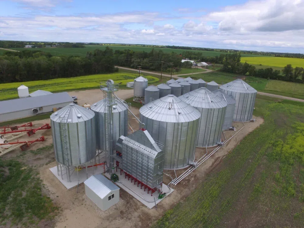 Aerial view of a modern grain storage facility with multiple corrugated steel grain bins, grain dryer, conveyors, and grain handling infrastructure surrounded by farmland.