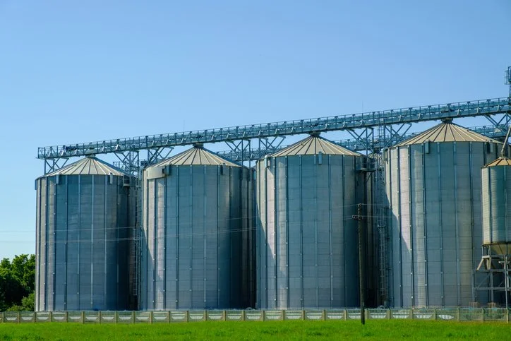 Large silver industrial silos in a row under a clear blue sky, illustrating where proper ventilation is required to prevent moisture migration in grain storage facilities.