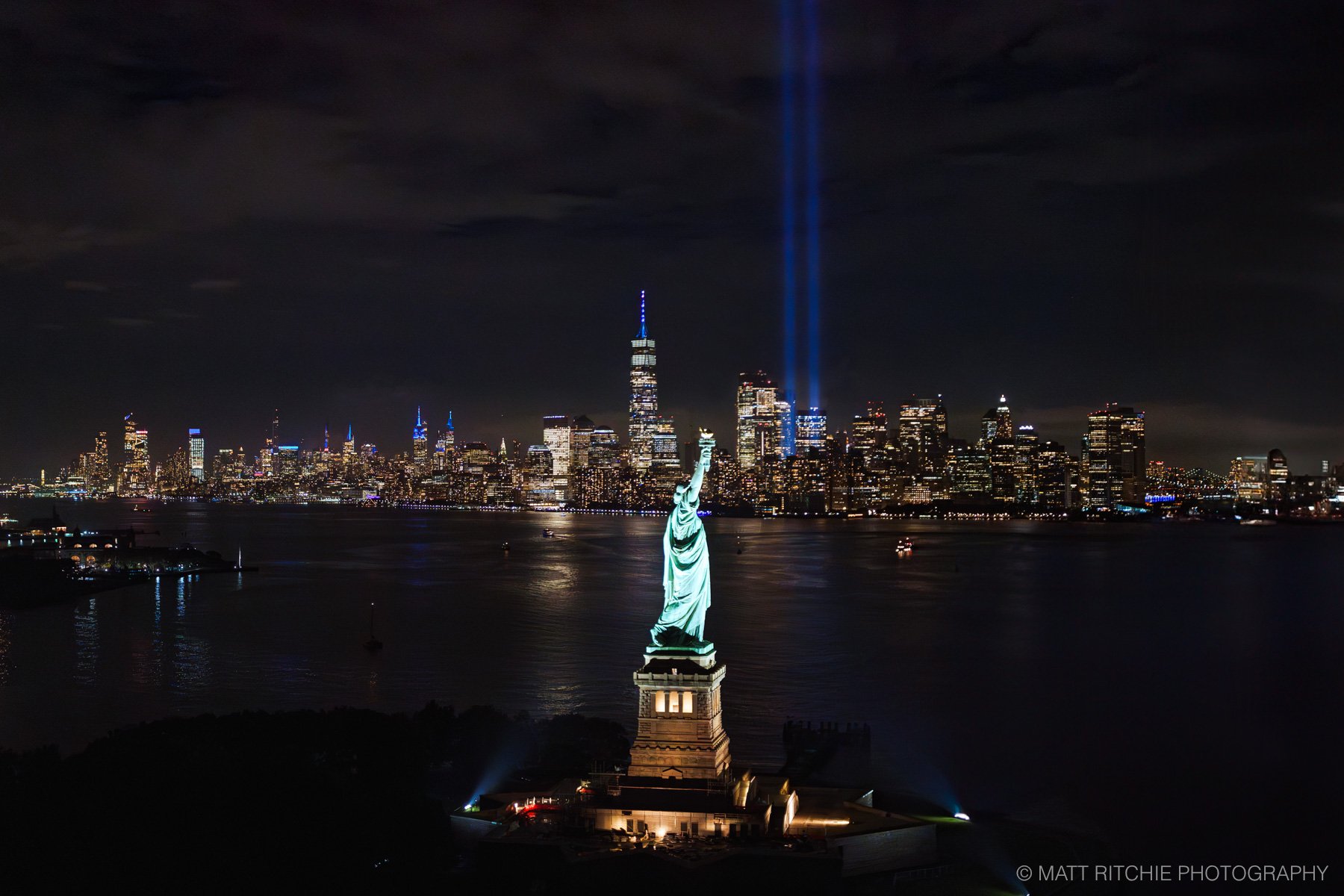 The Tribute in Light memorial illuminating the New York City skyline at night, with the Statue of Liberty in the foreground, photographed from a helicopter