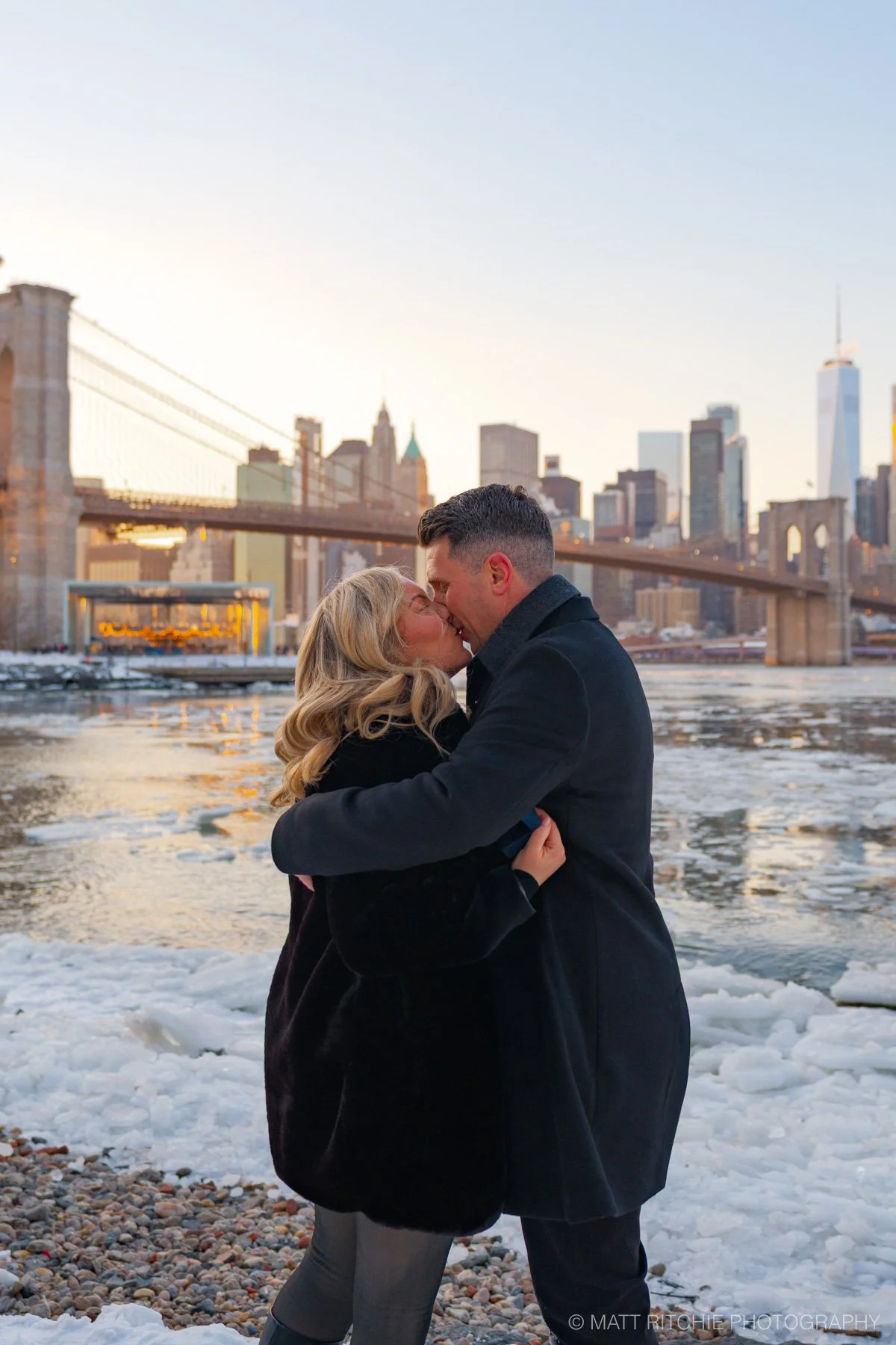 Winter marriage proposal in DUMBO Brooklyn with snow dusting the waterfront and the Manhattan Bridge in the background