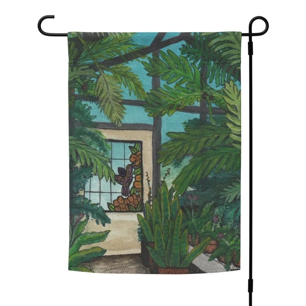 Garden flag featuring a greenhouse with ferns, cream colored door with stained glass of hummingbird and flowers. Bright blue sky shows through the windows.