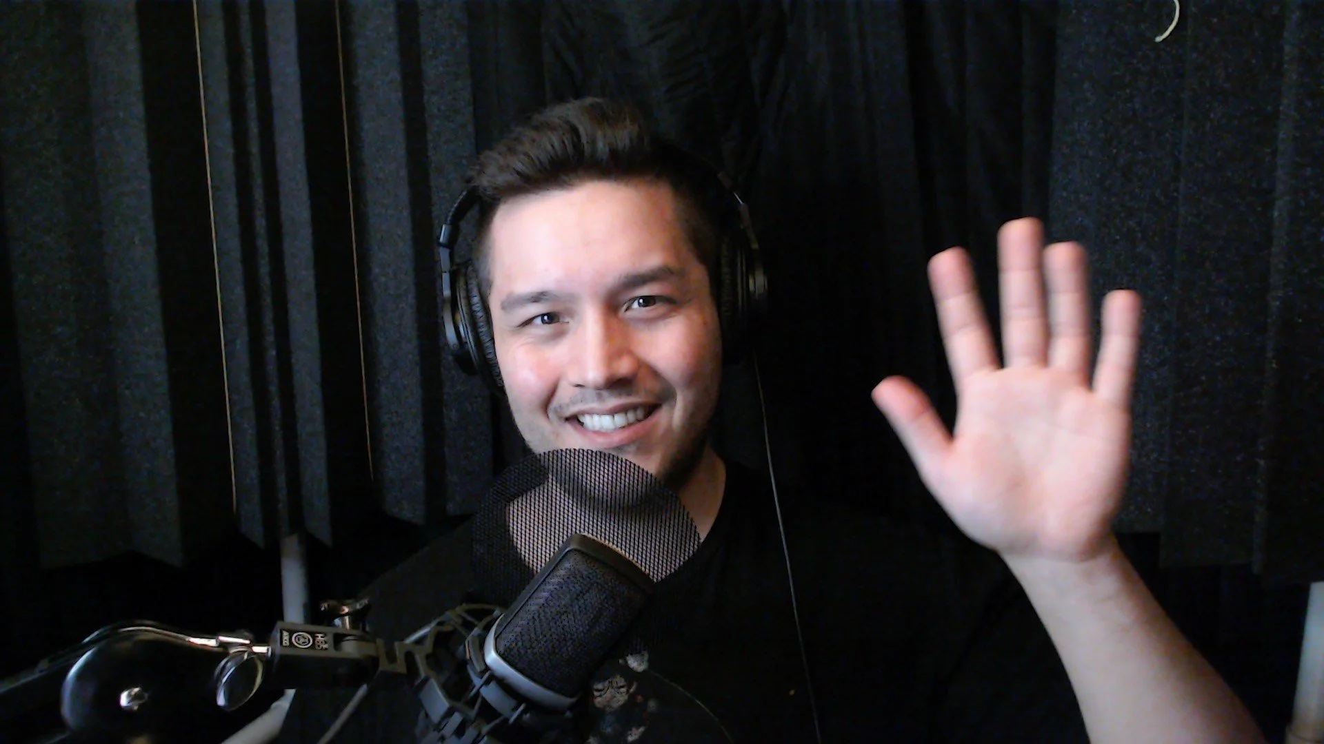 A young man with dark hair and a beard smiling and waving at the camera in a recording studio with soundproof black acoustic panels, wearing headphones, with a professional microphone in front of him.