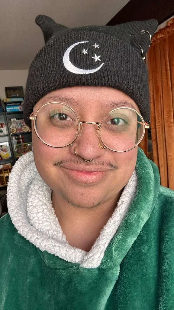 A self-taken photo of Felix Graves, a brown trans man with a thin mustache, several nose piercings, and wearing a black beanie and round, gold-framed glasses. He has a slight smile and is looking into the camera.