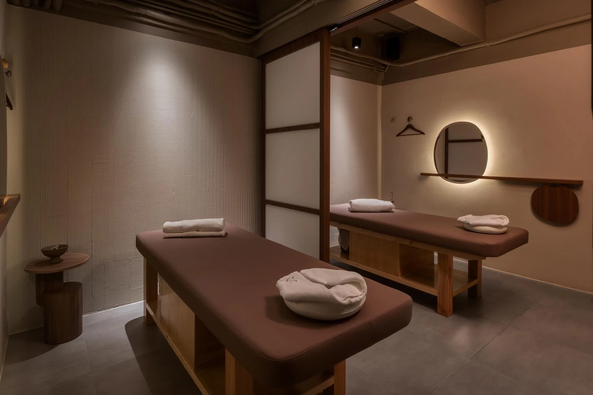 Two massage beds with rolled towels in a minimalist spa room with warm lighting and wooden accents.