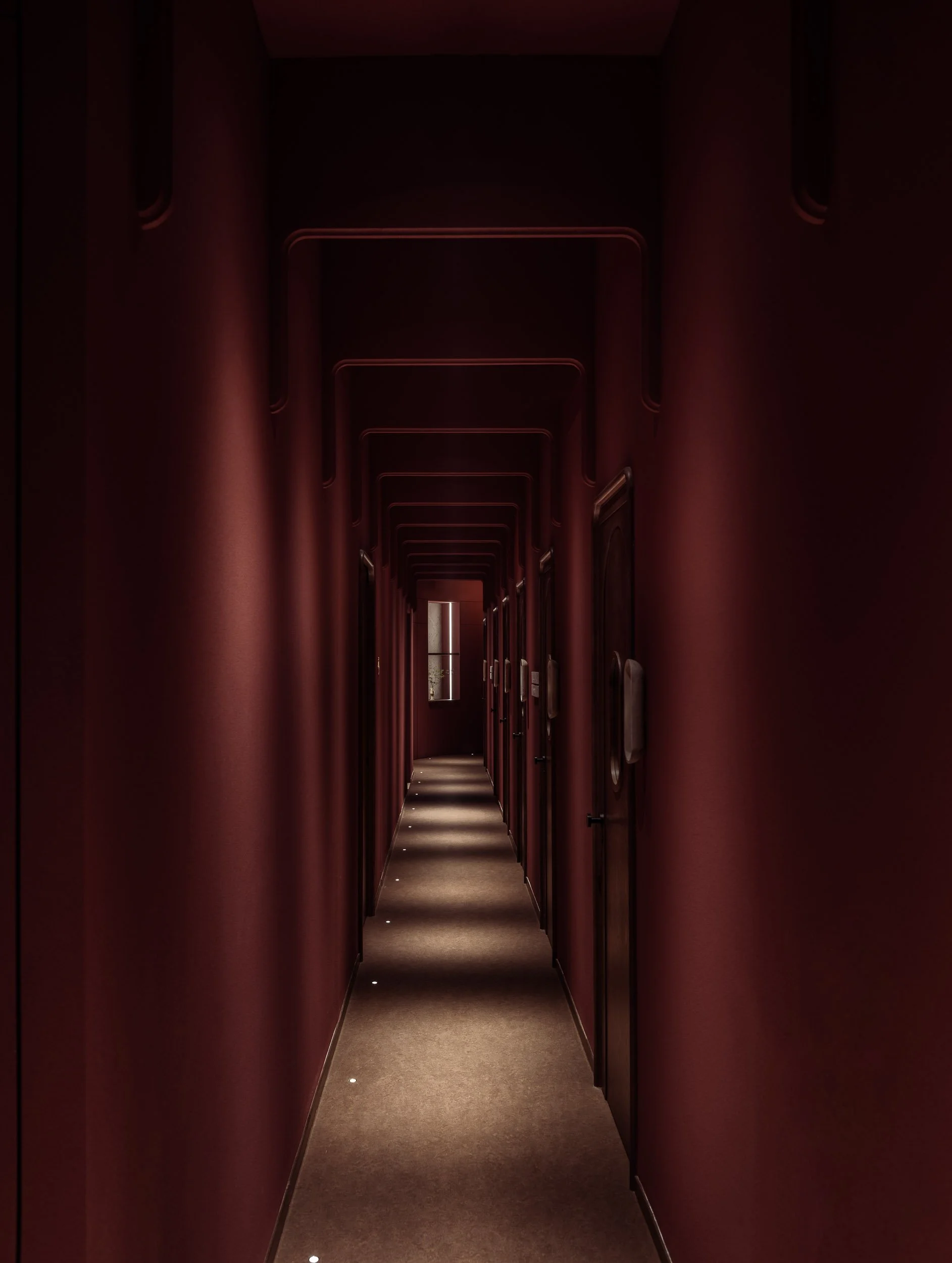Dimly lit narrow hallway with burgundy walls, a beige carpet, small ceiling lights, and multiple closed doors on each side.