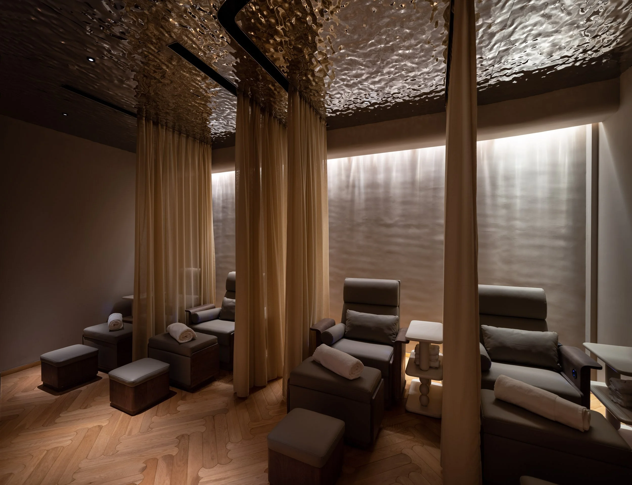 Spa or relaxation room with four gray lounge chairs, each with towel and pillow, divided by beige curtains, and soft lighting on textured beige wall.
