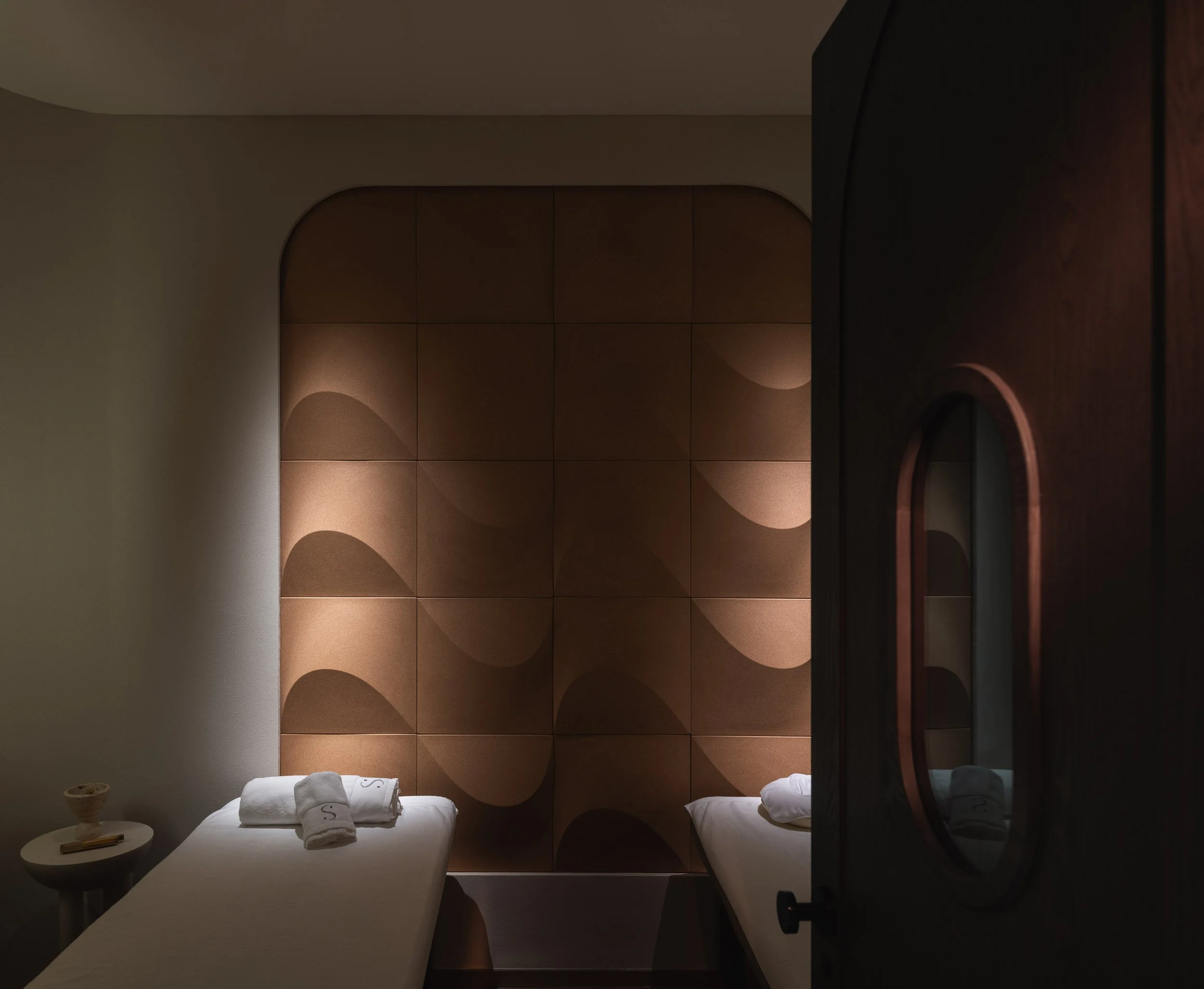 Two massage tables in a spa room with beige walls, a decorative textured wall panel, and a wooden cabinet with an oval glass window.