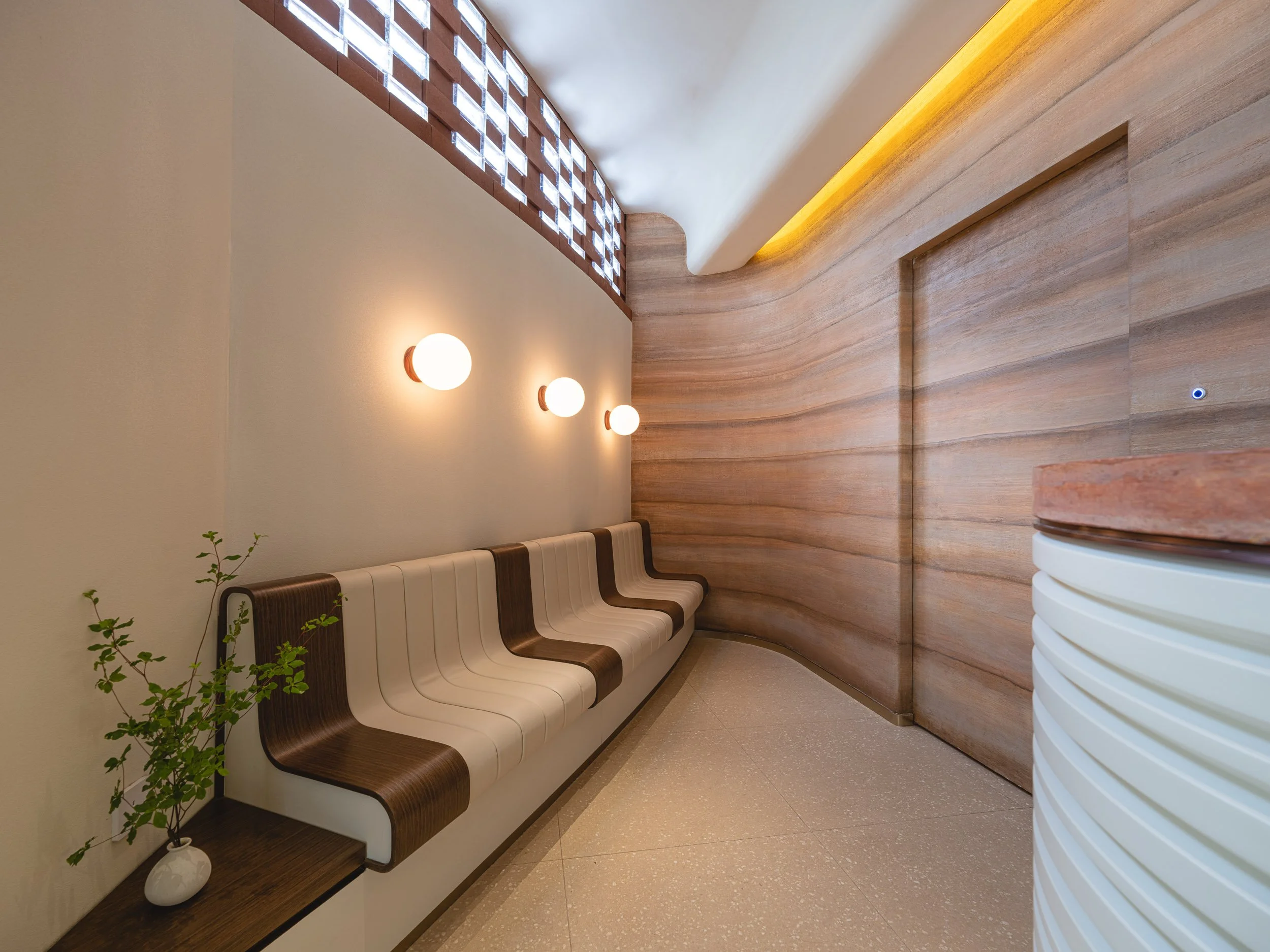 Modern waiting area with white and brown curved bench, small plant in white vase, wall mounted round lights, wooden panel wall, small window with lattice, and curved ceiling with yellow lighting.