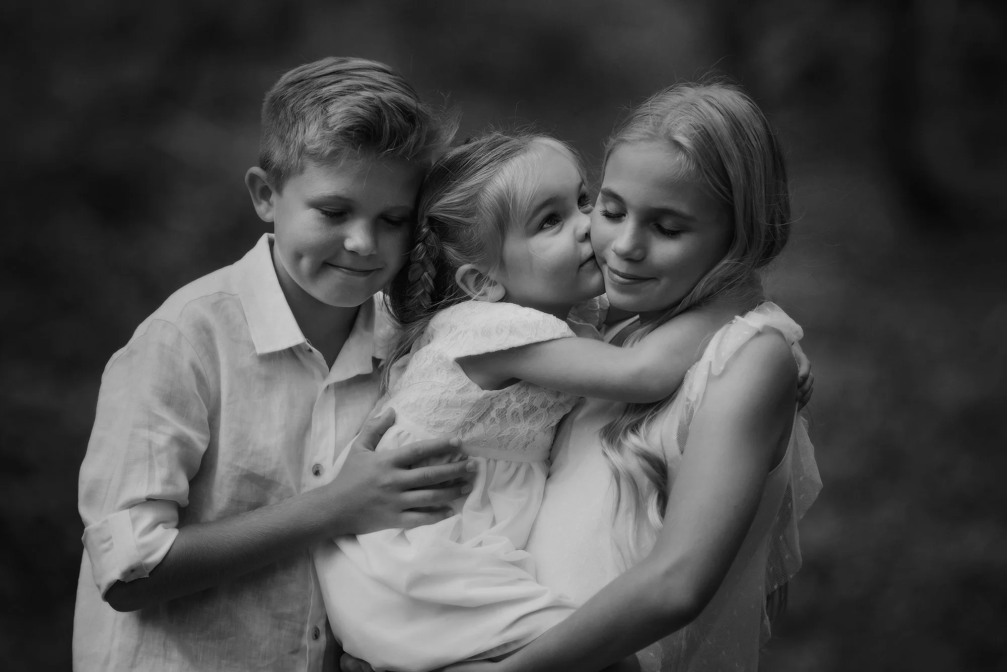 Black and white photo of three children hugging, with one girl kissing another girl's cheek and a boy smiling with eyes closed.