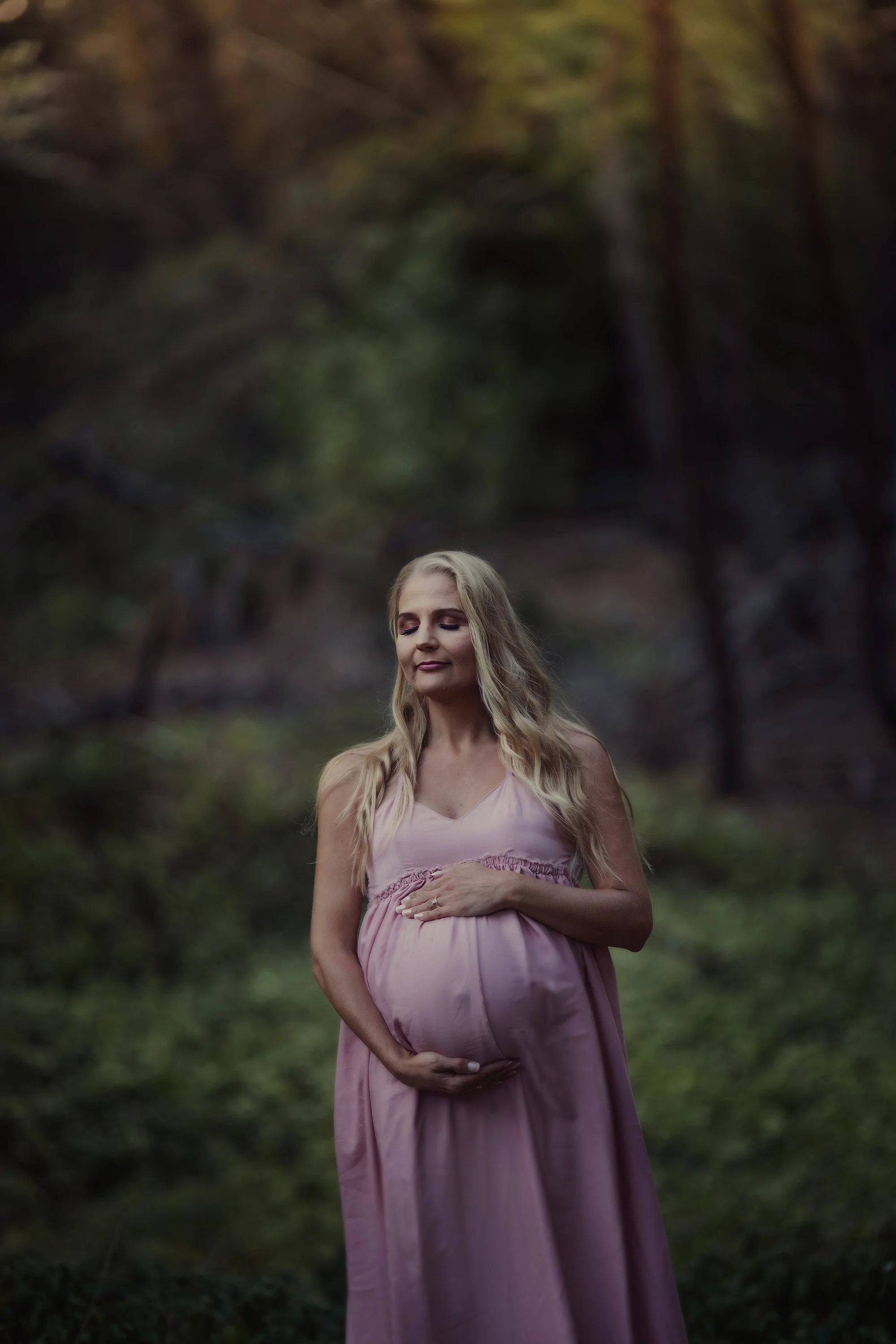 A pregnant woman in a pink dress standing outdoors in a wooded area, touching her belly with her eyes closed and a peaceful expression.