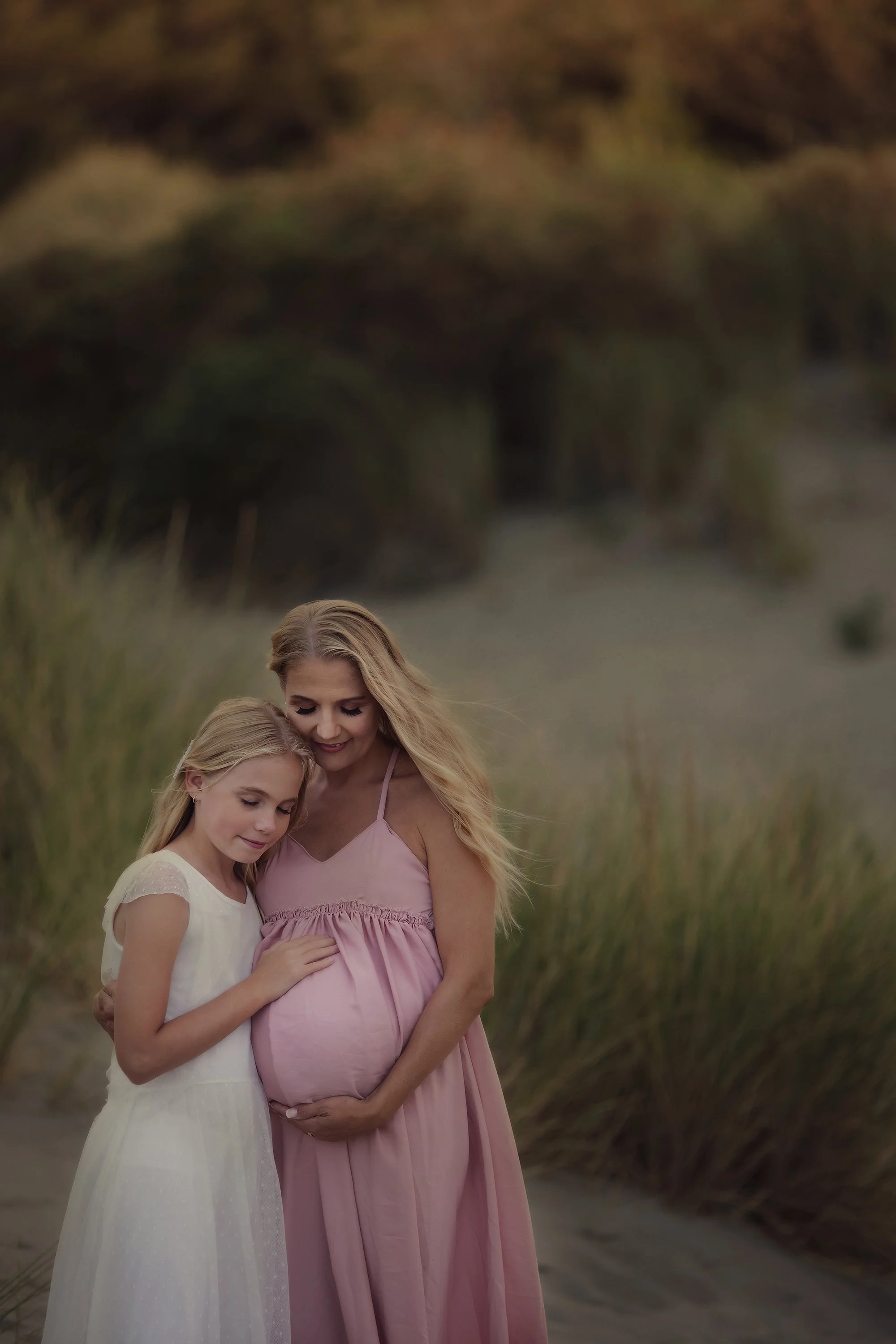 A woman and two girls standing outdoors in a grassy area, with the woman pregnant, all with their heads bowed and eyes closed, sharing a tender moment.