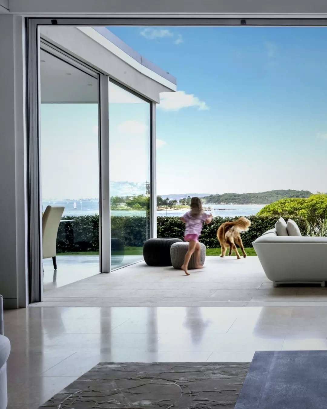 Set beneath a broad roofline, the terrace extends the living area, furnished simply with low seating and soft textures. The composition of floor, ceiling and glazing forms a clear edge to the room, allowing the open air, greenery and distant water vi