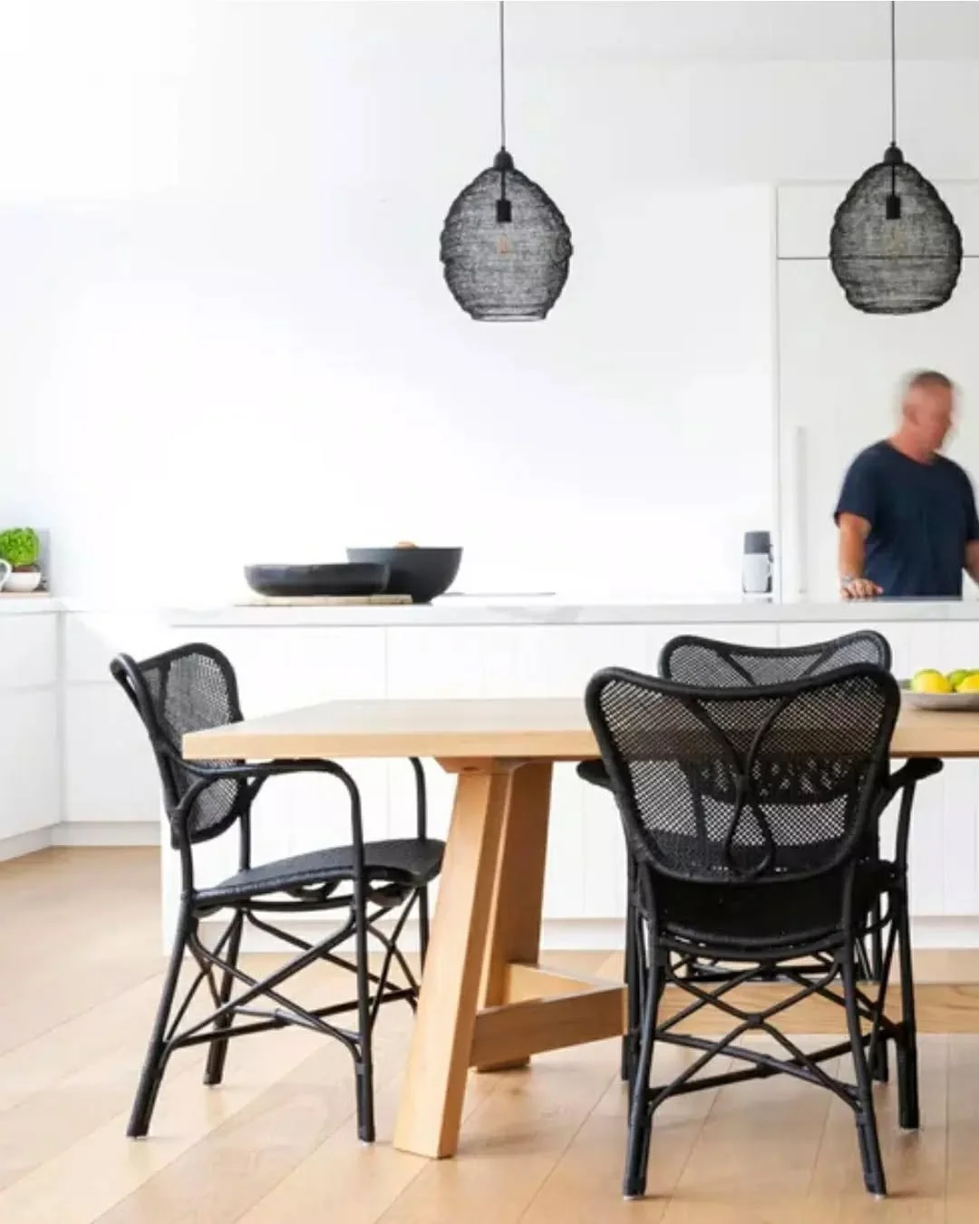 Defined by a restrained material palette and generous natural light, the kitchen and dining area centres around a large timber table that anchors the space. Pale oak flooring and crisp white cabinetry create a calm, open backdrop, while black woven d