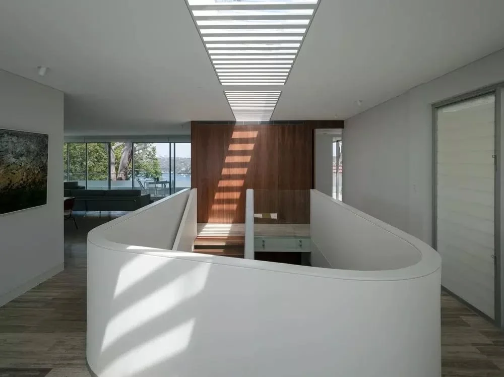 A central stair is introduced as a defining element within the upper level, formed by a curved balustrade that shapes the void below. The smooth white form sits alongside a timber wall, where material contrast and precise detailing give the stair a s