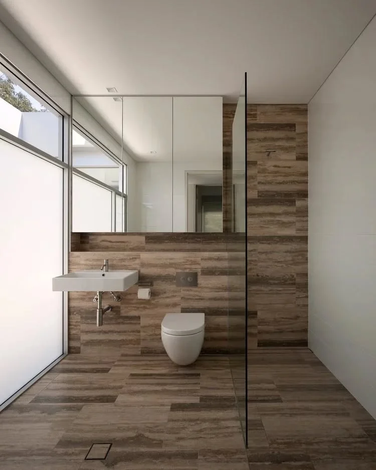 The bathroom layout is organised around a clear division between wet and dry areas, with a frameless glass screen maintaining openness within the space. Large-format stone tiles extend across the floor and walls, establishing a consistent material la