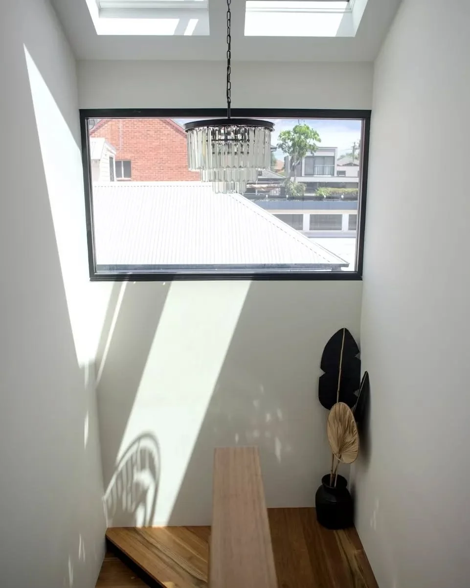 A double-height stair void draws natural light deep into the interior, with twin skylights positioned above a large picture window. Light shifts across the walls and timber stair throughout the day, while dark window framing contrasts against crisp w