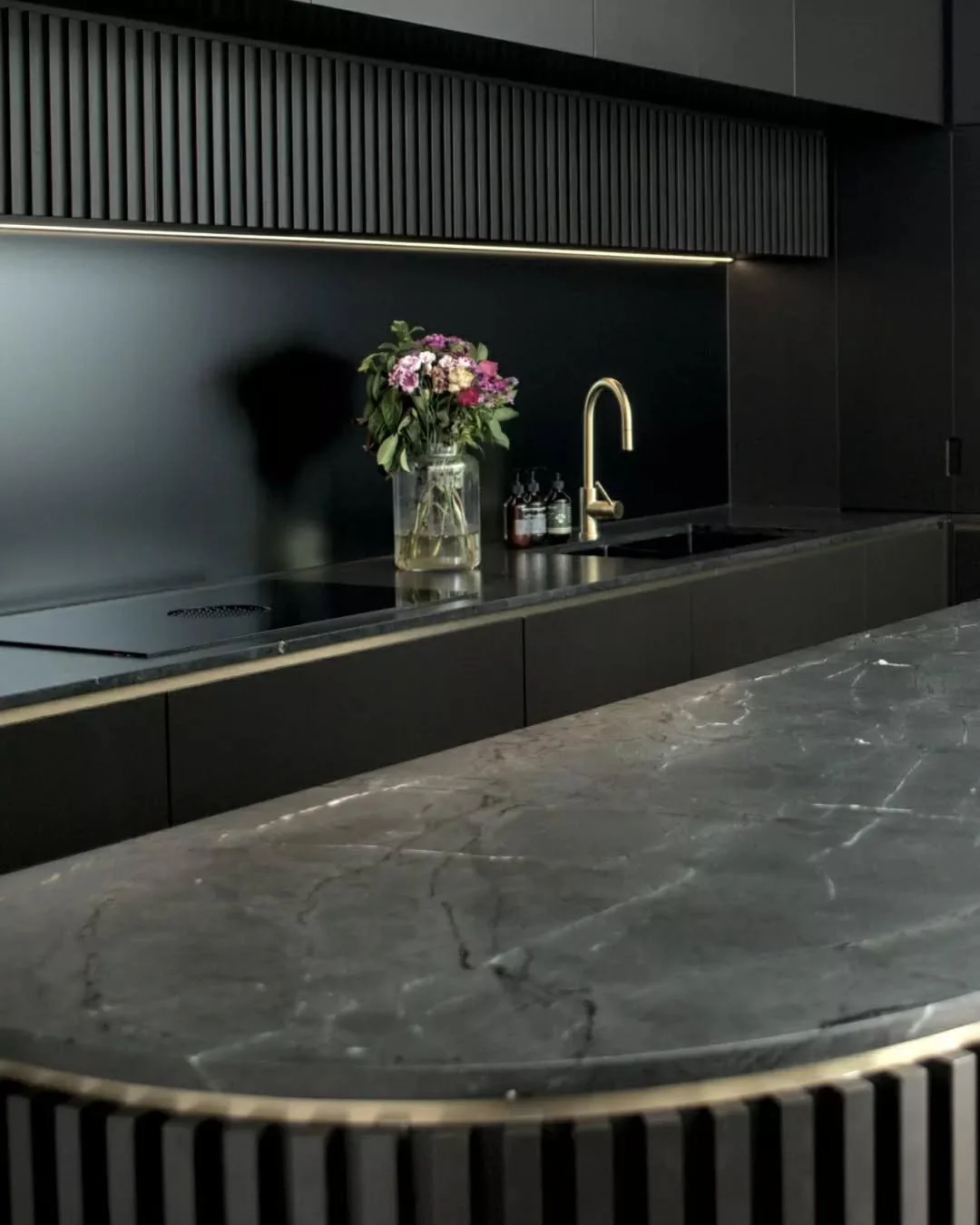 Fluted joinery forms a continuous overhead feature, introducing texture against dark cabinetry below. A slimline linear light runs beneath, softly illuminating the splashback. Integrated cooktop and sink maintain a minimal surface, allowing materiali
