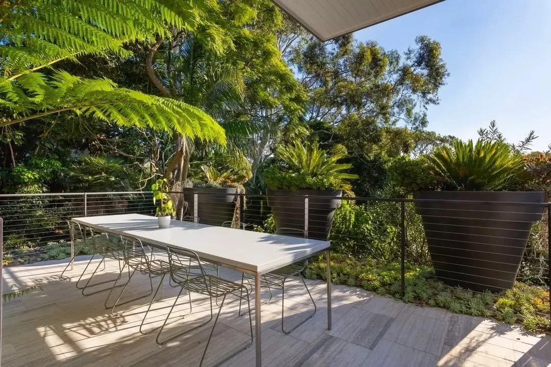 Large sliding glass doors open to a tiled terrace framed by slim steel posts, extending the living areas outward. The covered terrace sits beneath a deep overhang, with a lounge setting arranged to overlook surrounding foliage and a dining area posit