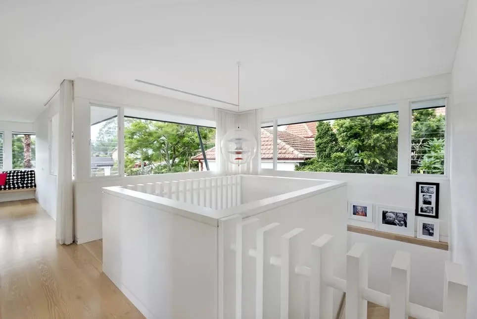 Corner windows wrap the upper level, bringing daylight across the stair landing and framing outward views. A white balustrade encloses the stair opening, forming a sculptural element that anchors the space, while pale timber flooring adds warmth bene