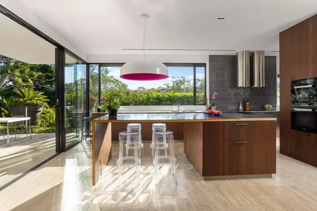 A large picture window stretches across the kitchen, drawing light onto the stainless steel benchtop. Warm timber cabinetry anchors the space, while the island introduces a contrasting surface with integrated seating below. Clear stools maintain an o