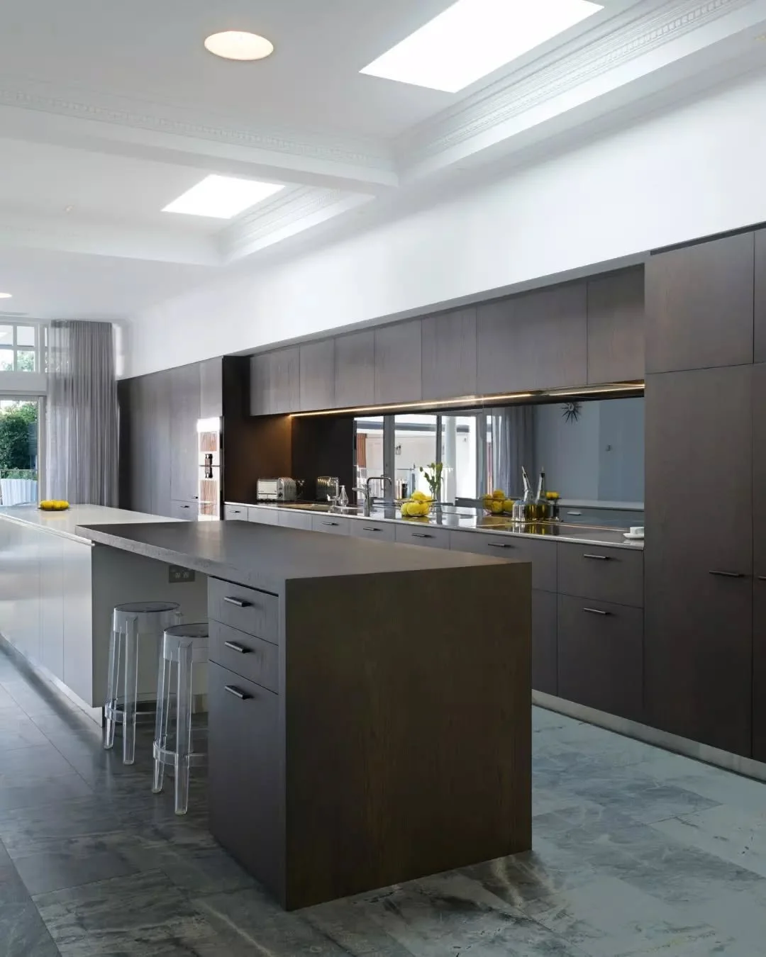 Dark timber joinery runs the length of the kitchen, introducing contrast beneath the elevated ceiling and skylights above. A long stone benchtop and integrated Gaggenau appliances, selected for their sharp design, maintain a clean and continuous fini