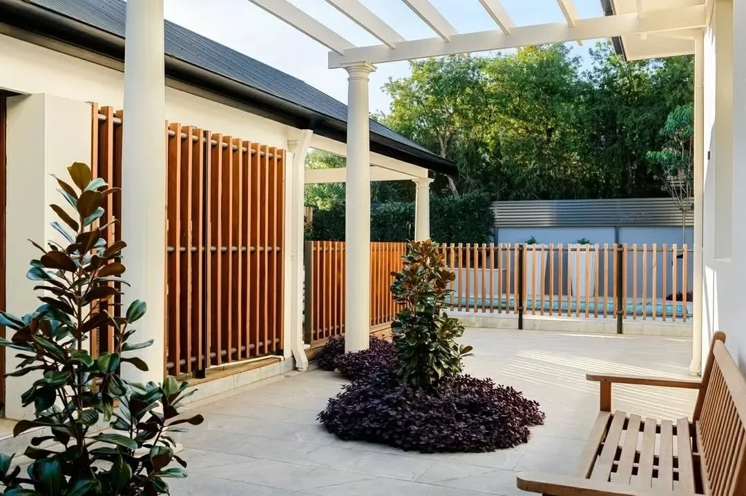 Granite paving extends through a pergola-covered walkway linking the side entrance to the rear garden and pool area. Classical columns and overhead beams frame the outdoor passage, while soft landscaping introduces texture and greenery along the edge