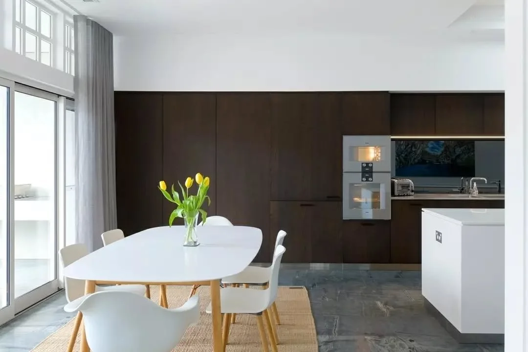Expansive sliding doors open the dining and kitchen areas toward the terrace and pool, with highlight windows above retaining the detailing of the original home. Stone flooring runs throughout the interior, while the outdoor areas remain framed withi
