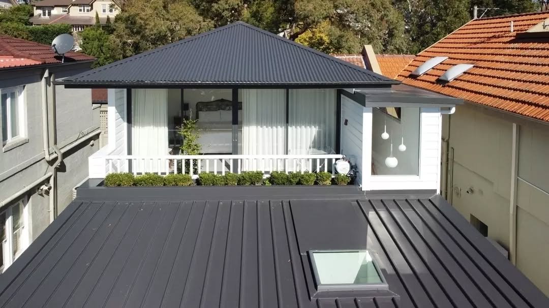A new upper-level addition introduces a simple form with white cladding, dark roofing and a monochromatic exterior finish. Sliding doors extend onto a narrow balcony framed by low landscaping and white balustrades.

Positioned above the existing stru