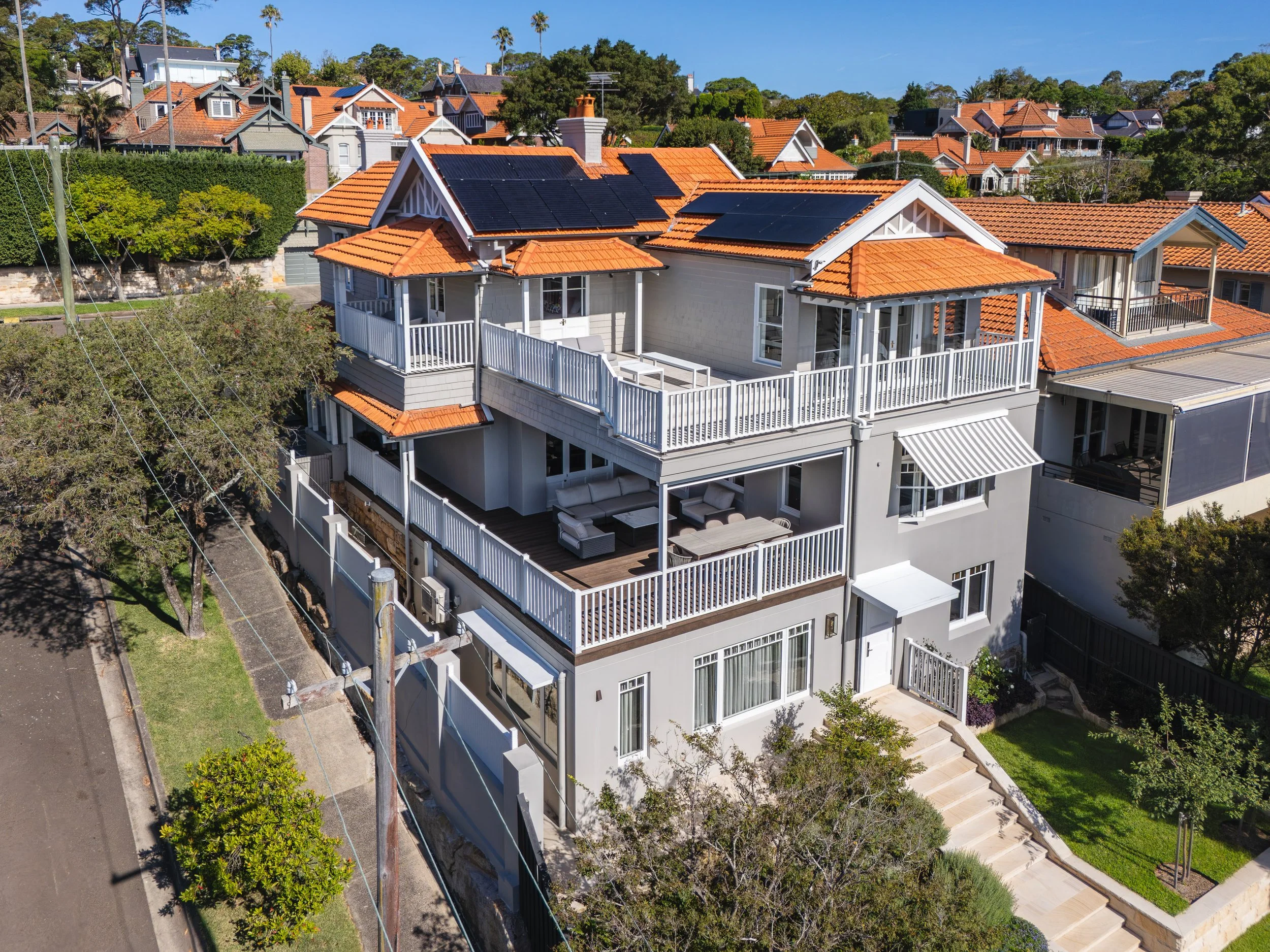 A modern multi-level house with gray exterior walls and orange tiled roofs, featuring several balconies with white railings and solar panels on the roofs, surrounded by trees and neighboring houses with similar architectural styles.
