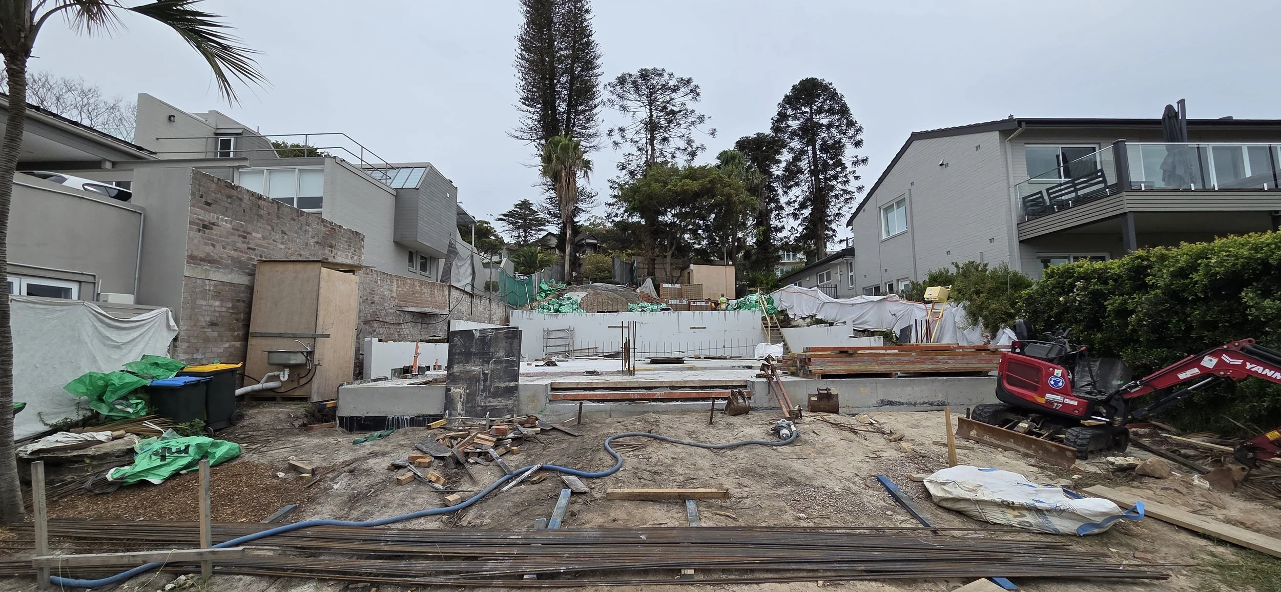 Construction site with building foundation, construction equipment, and neighboring houses in the background.