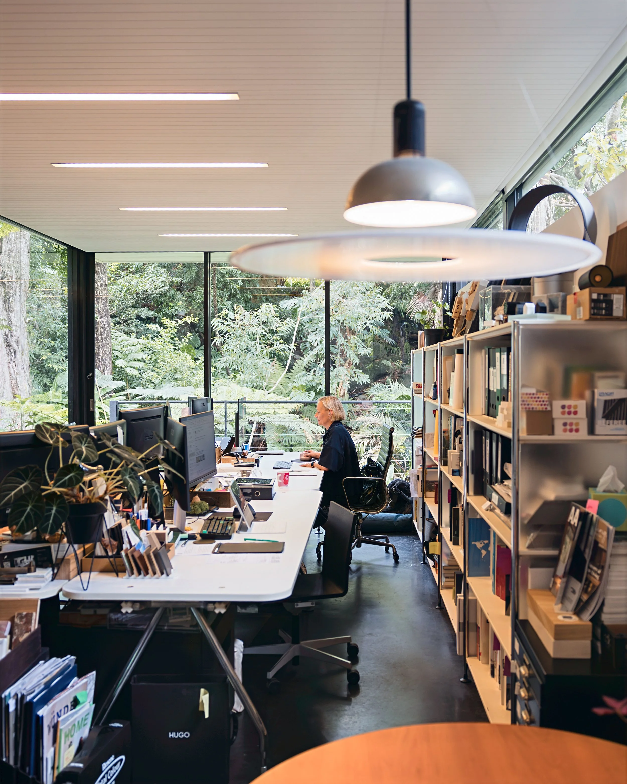 Architect working at desk in a modern office with large windows and lush greenery outside, surrounded by shelves with books, models and materials