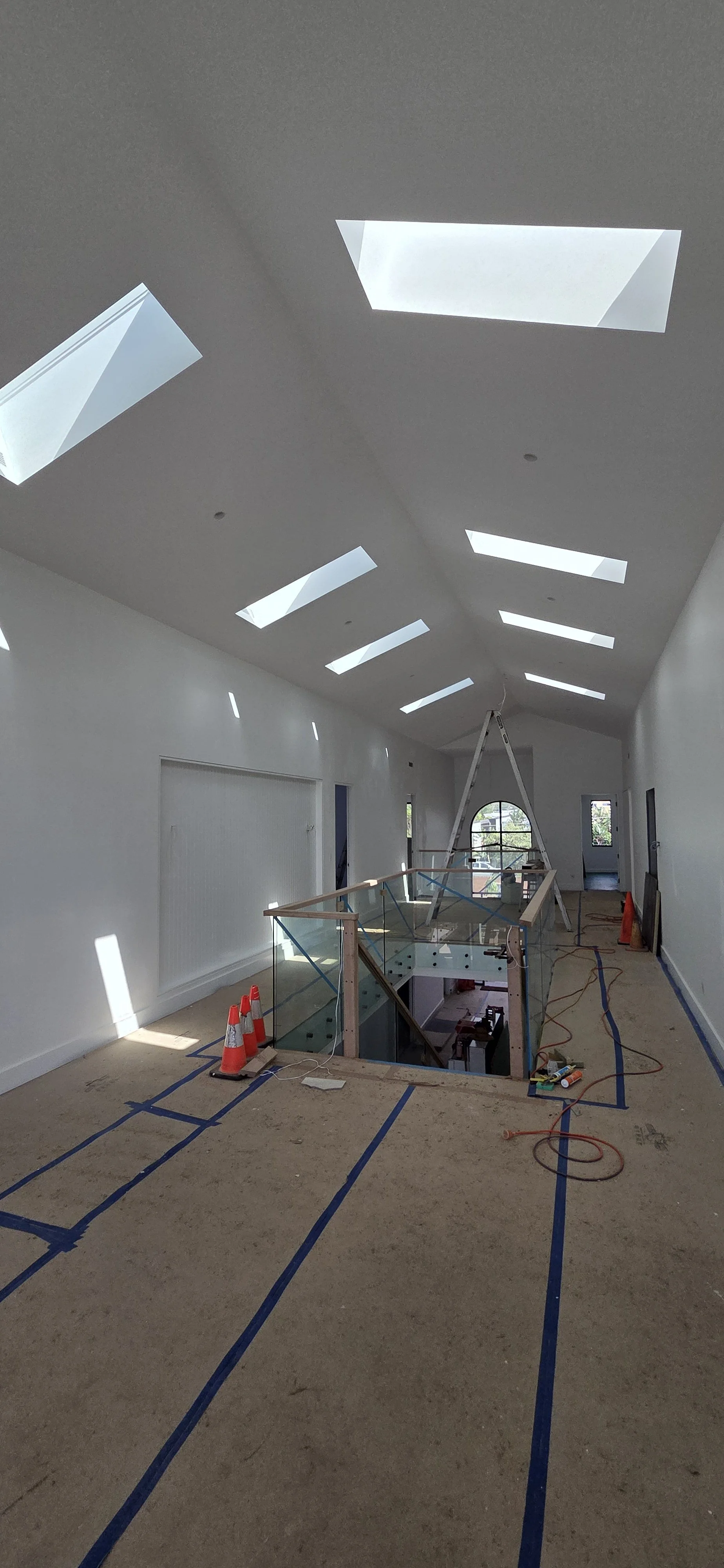 Interior of a building under construction, with skylights in the ceiling, a glass railing around an open staircase, construction cones, and tools on the floor.