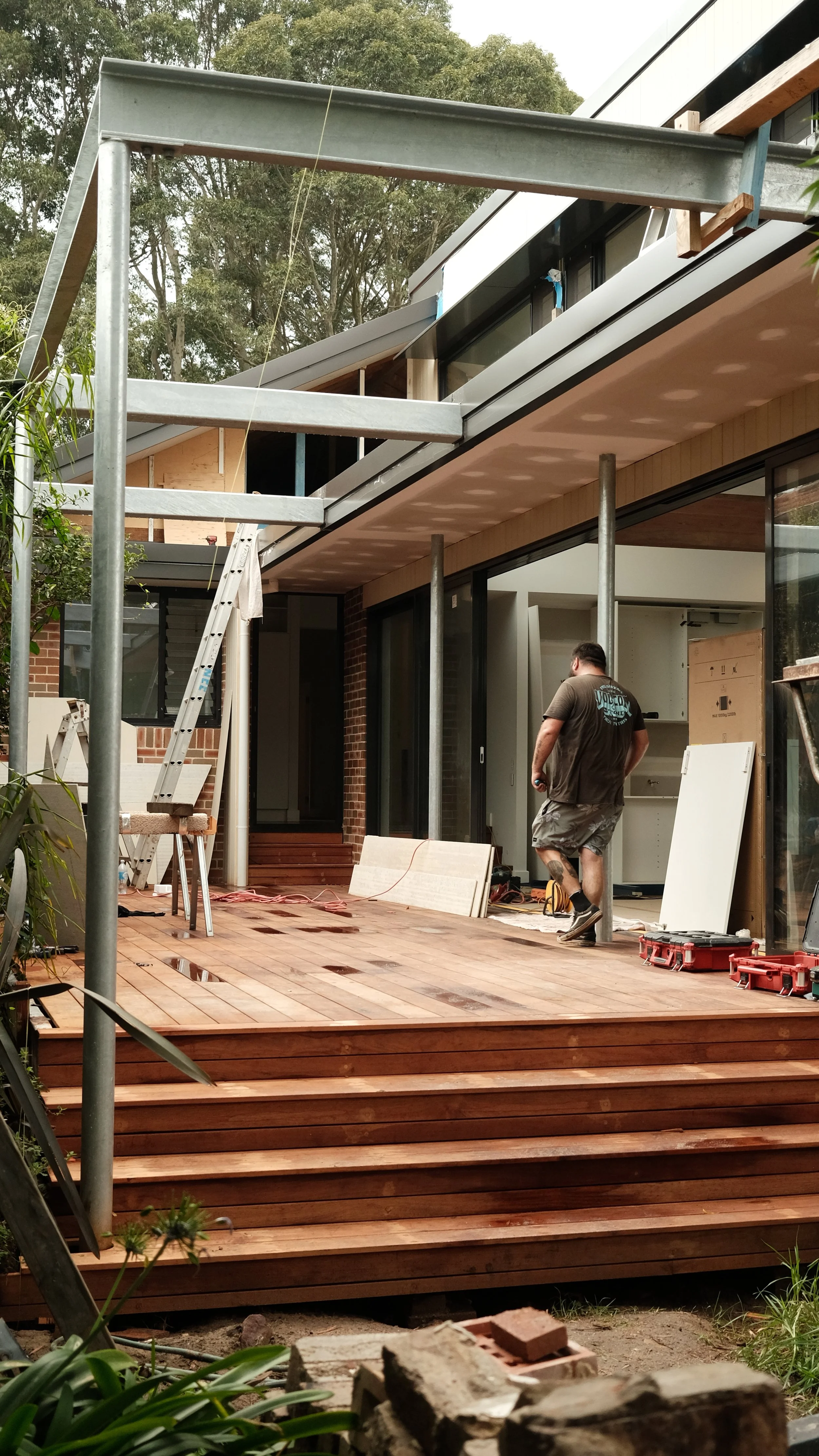 A worker constructing a wooden deck outside a modern house with large sliding glass doors, with construction materials and tools around.