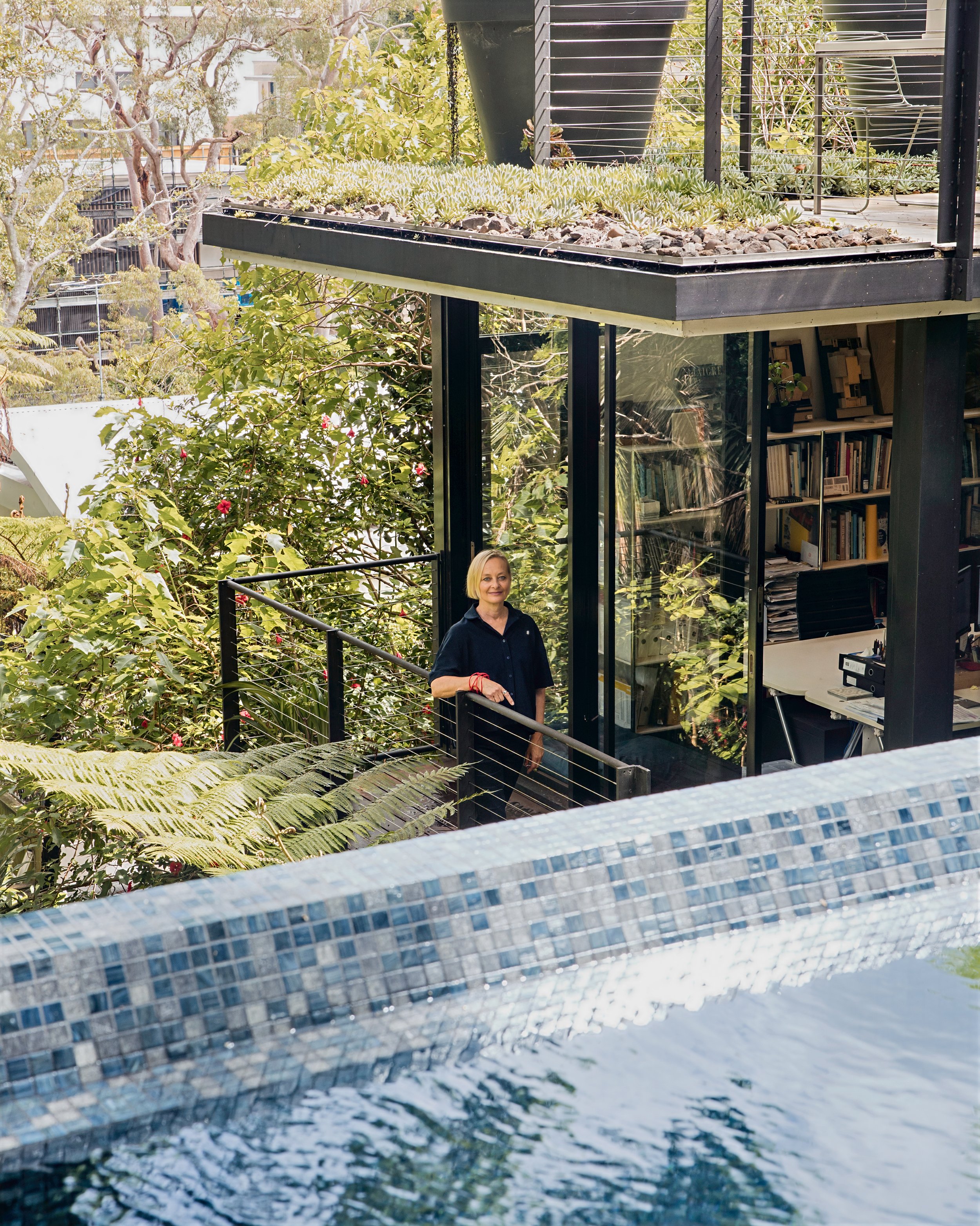 A woman smiling and standing on a balcony surrounded by lush greenery and plants, with a modern glass and metal structure and a bookshelf visible behind her.