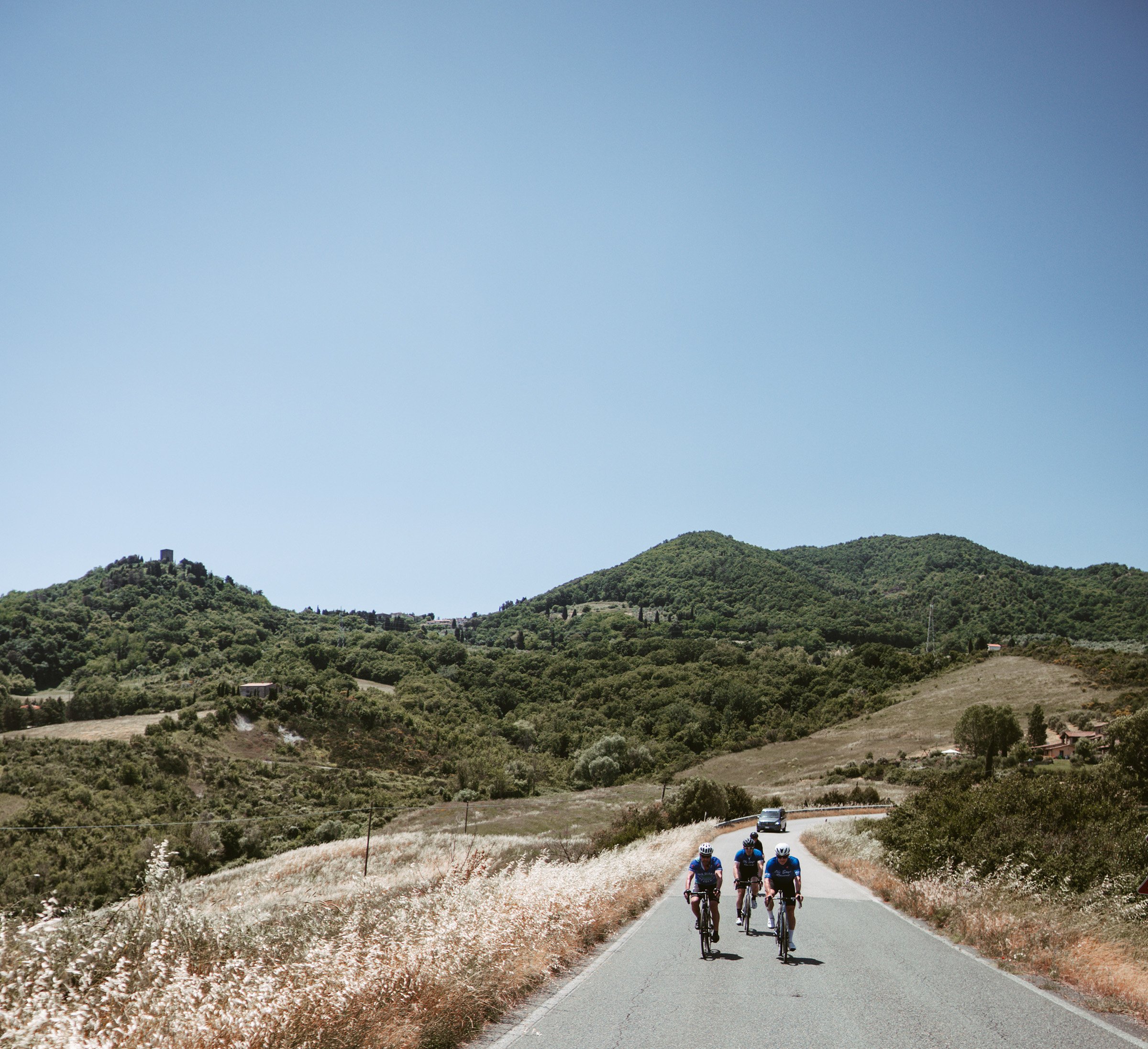 Three cyclists riding on a rural road through scenic hills with a clear blue sky.