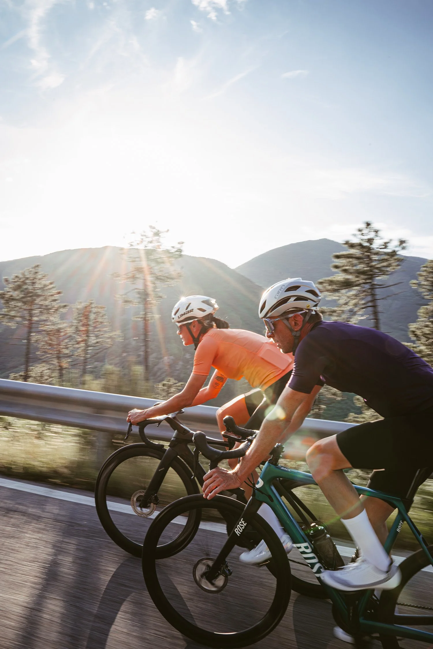 Two cyclists riding on a mountain road during sunset with trees and mountains in the background.