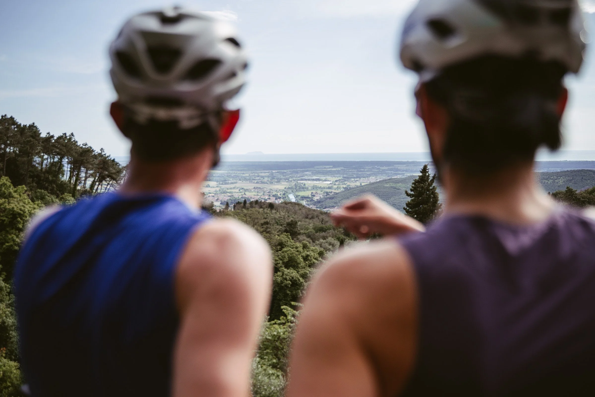 Two people wearing biking helmets and sleeveless shirts looking out over a landscape of trees, hills, and water in the distance.