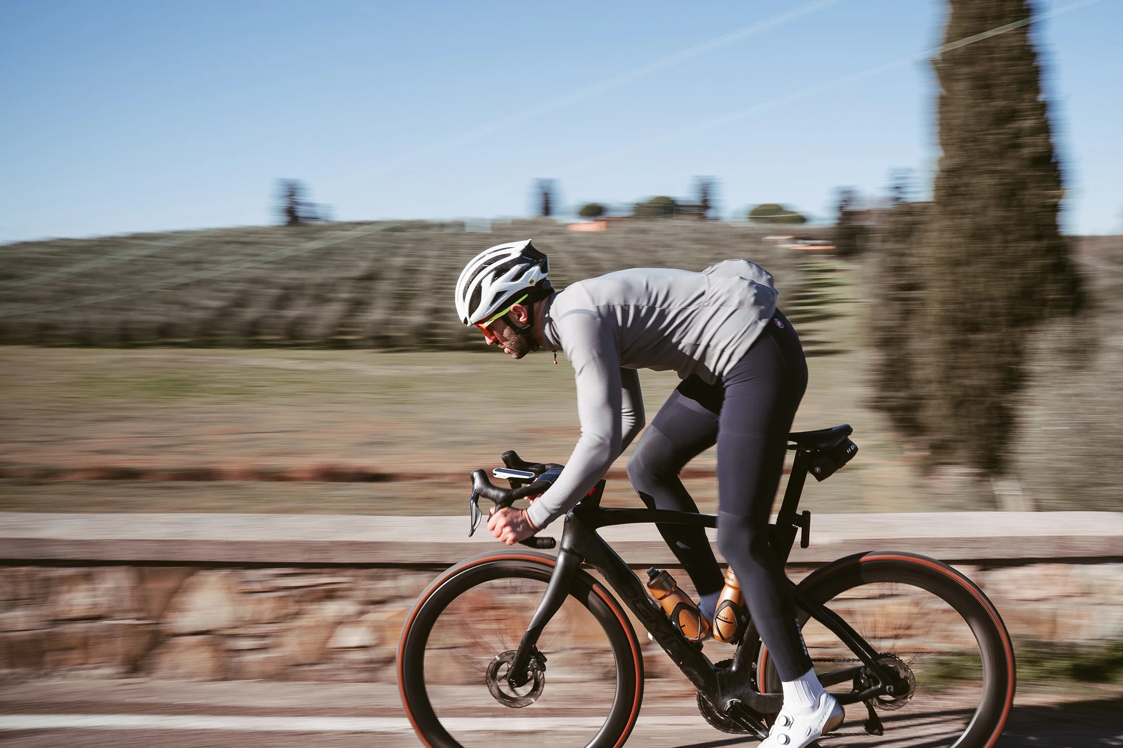 A man cycling outdoors on a mountain bike wearing a white helmet, sunglasses, a gray jacket, black pants, and white cycling shoes with red accents.