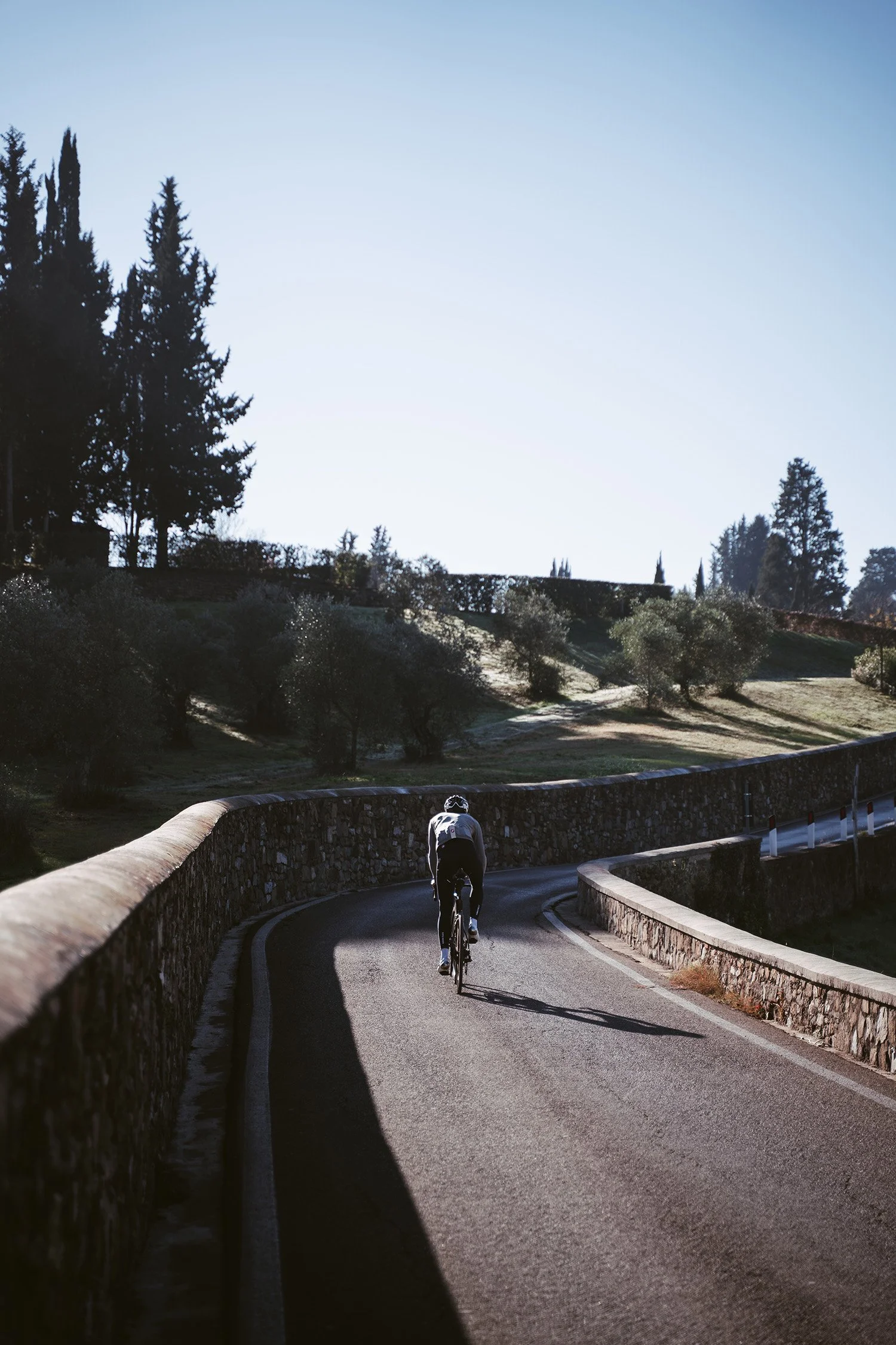 A cyclist riding on a winding paved road with stone walls on both sides, surrounded by trees and hilly terrain under a clear sky.