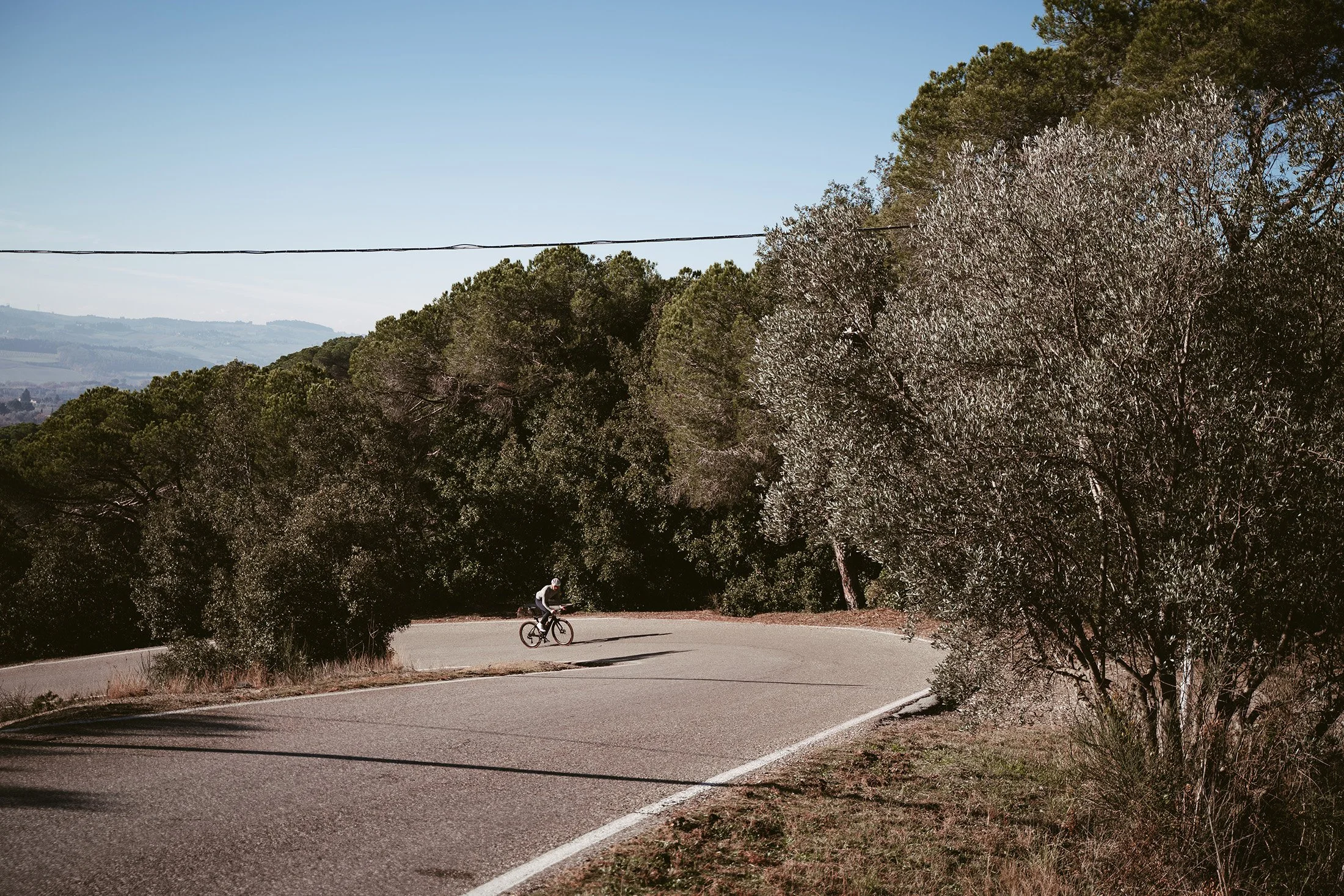 A person riding a bicycle on a winding mountain road with trees on both sides and hills in the background on a sunny day.