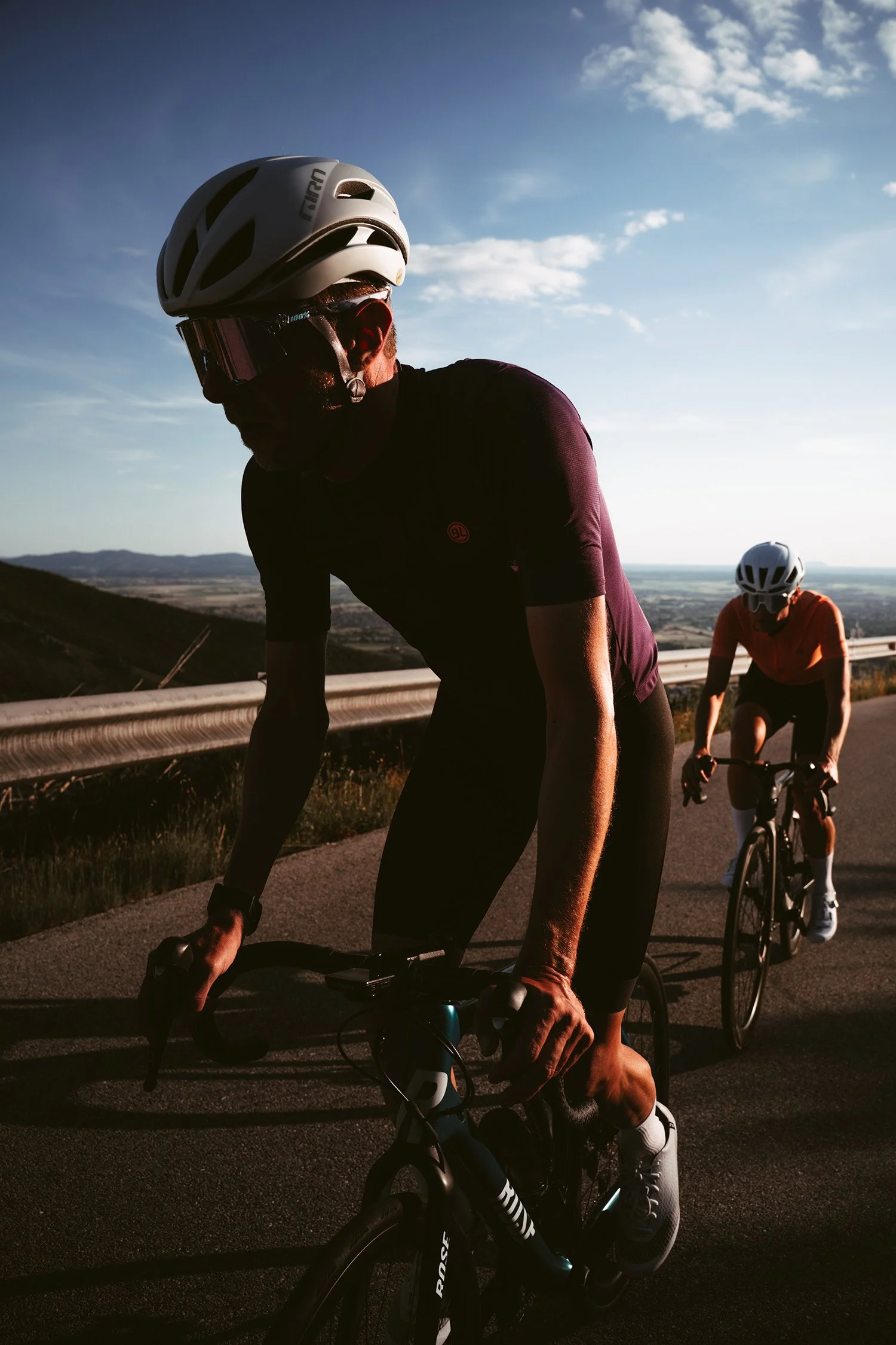 Two cyclists riding on a mountain road during sunset, wearing helmets and athletic clothing, with scenic landscape and clear sky in the background.