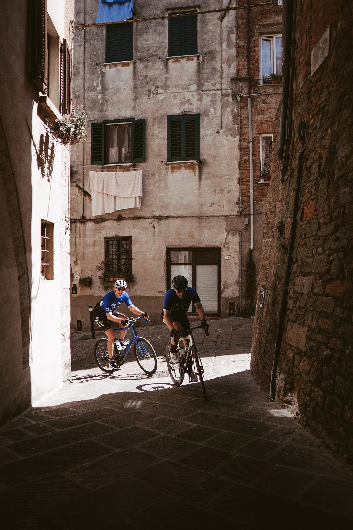 Two cyclists riding bikes through a narrow alleyway in a historic European town. The alley is made of stone and surrounded by old brick and stucco buildings with green shutters and hanging laundry.