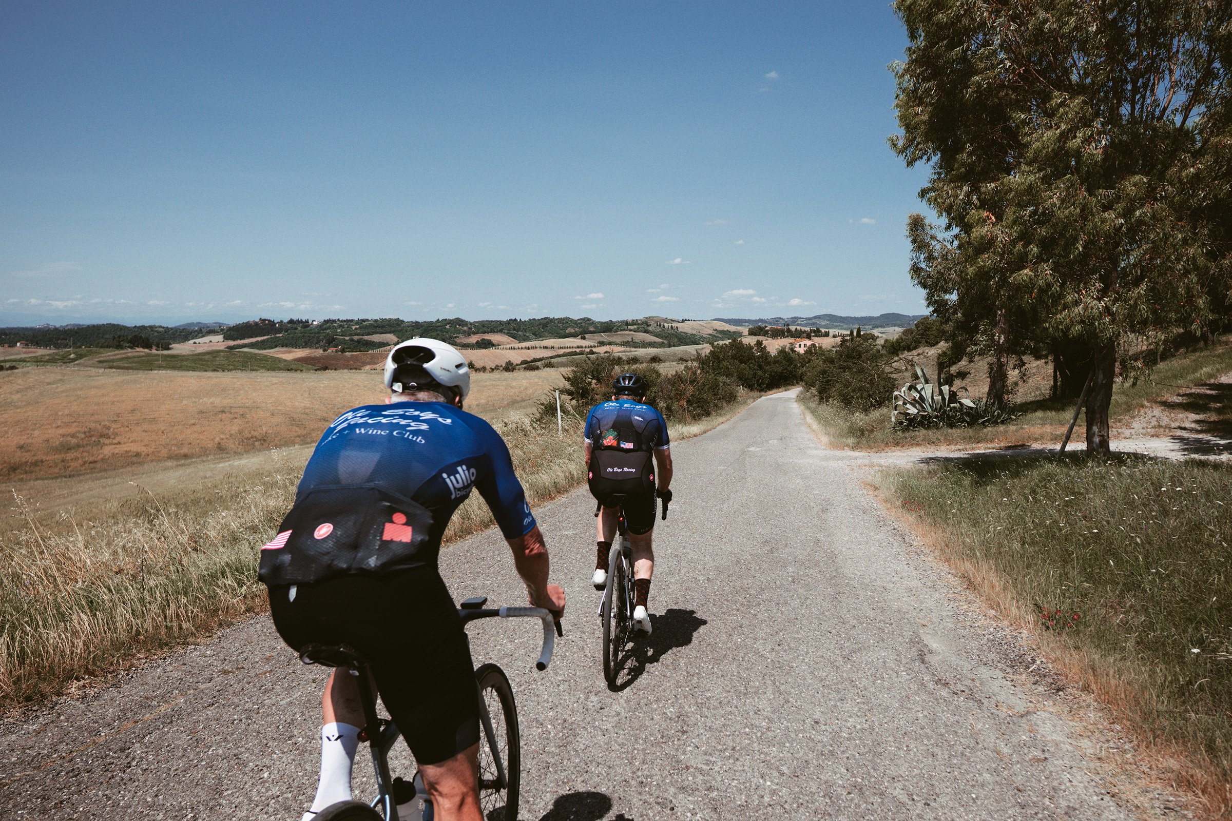 Three cyclists riding on a gravel road through a rural landscape with fields and trees under a blue sky.