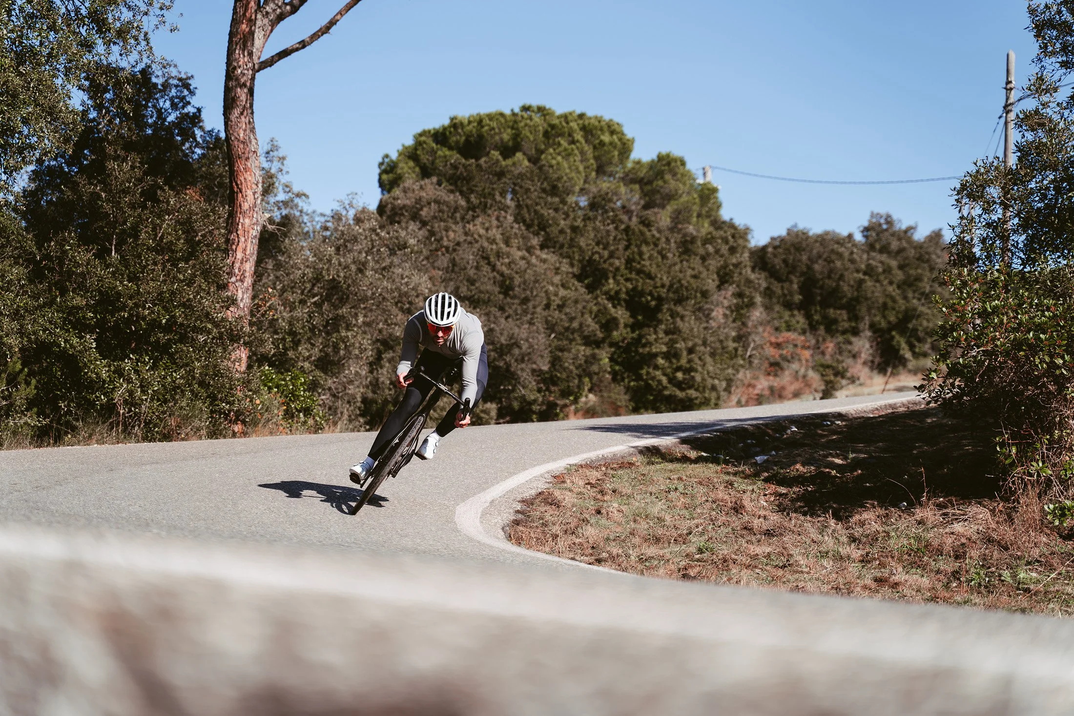A cyclist in a gray and black outfit riding a bike on a winding road through a wooded area with trees and bushes under a clear blue sky.