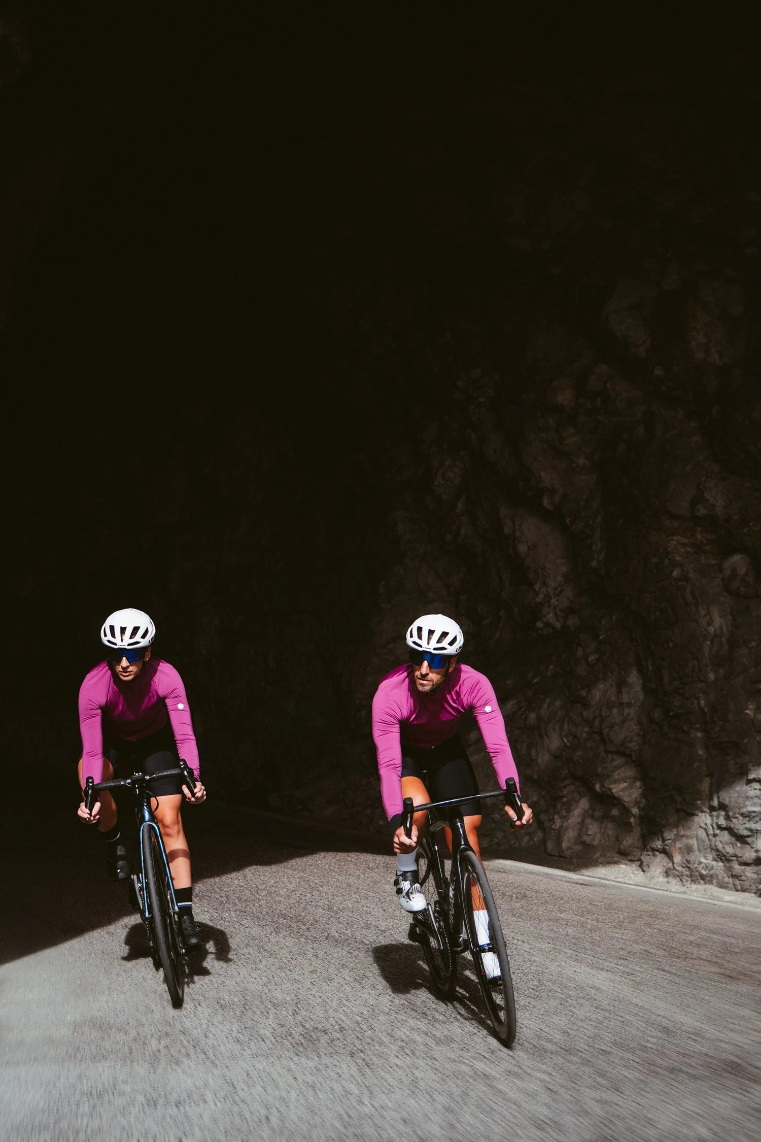 Two cyclists wearing white helmets and purple long-sleeve shirts riding on a road through a dark tunnel with rocky walls.